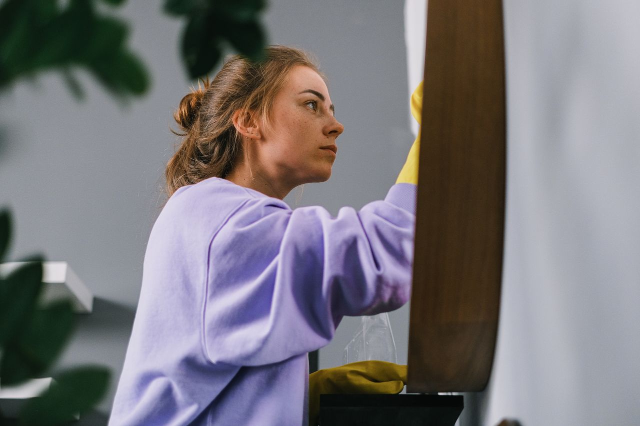 Side view of a young, concentrated woman in latex gloves standing and cleaning a mirror in a bright room during the day